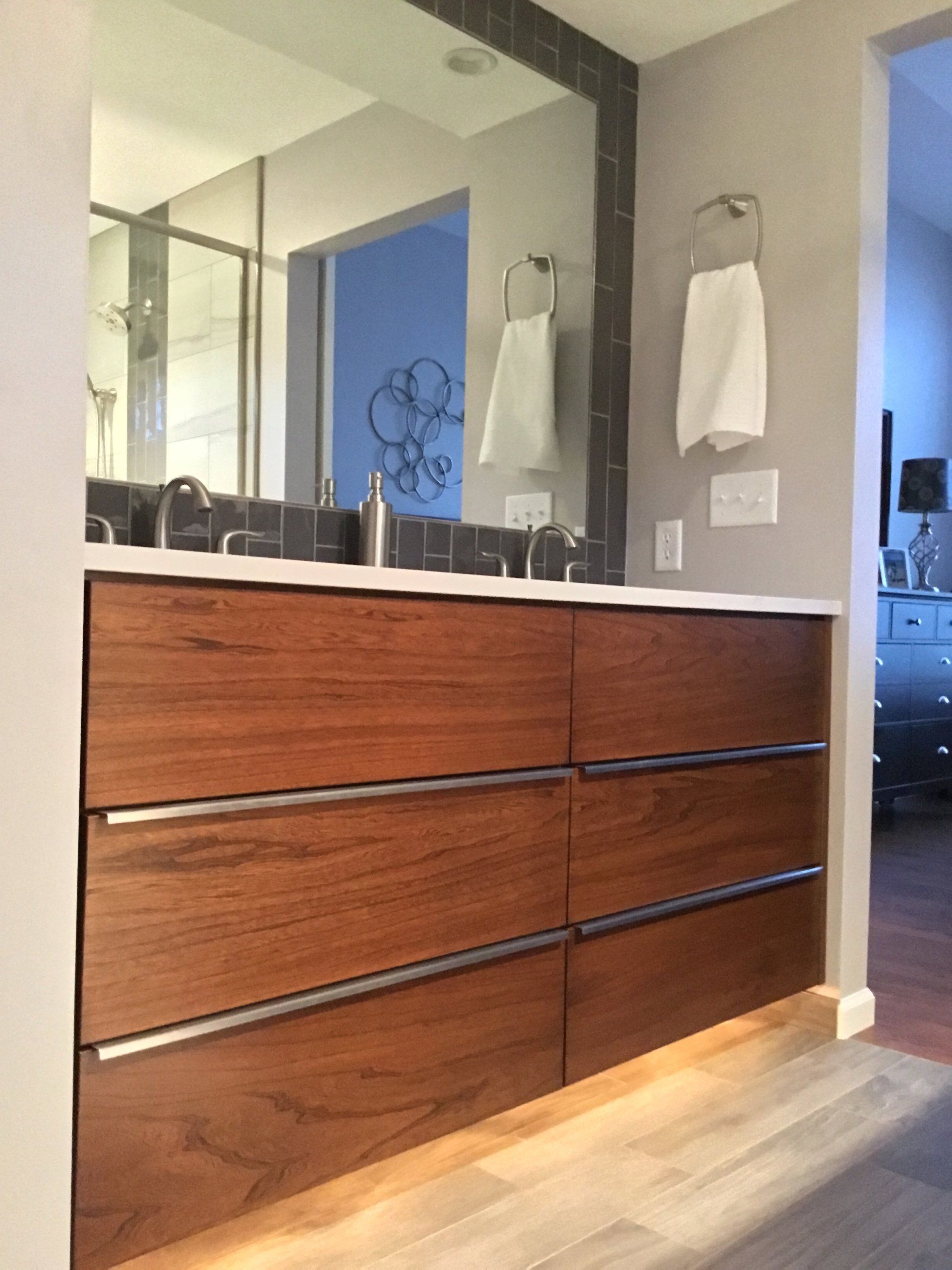 Modern bathroom vanity with wood drawers, white countertop, mirror, and towels on a gray wall.