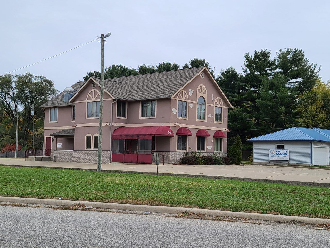 Two-story pink house with red awnings beside a small blue-roofed building and grassy roadside.