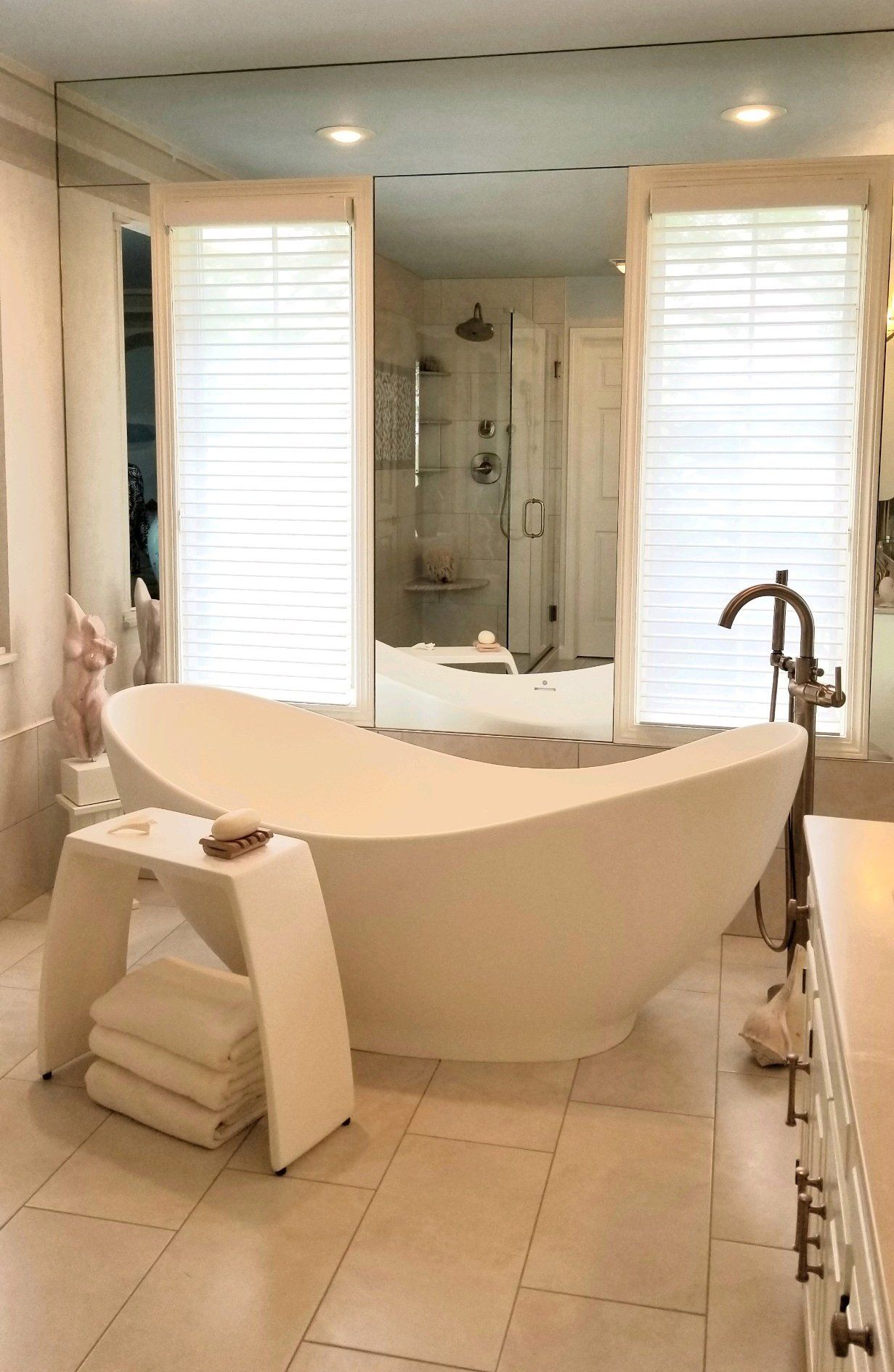 Bright bathroom with a white freestanding tub, large mirror, tiled floor, and shower visible in back.