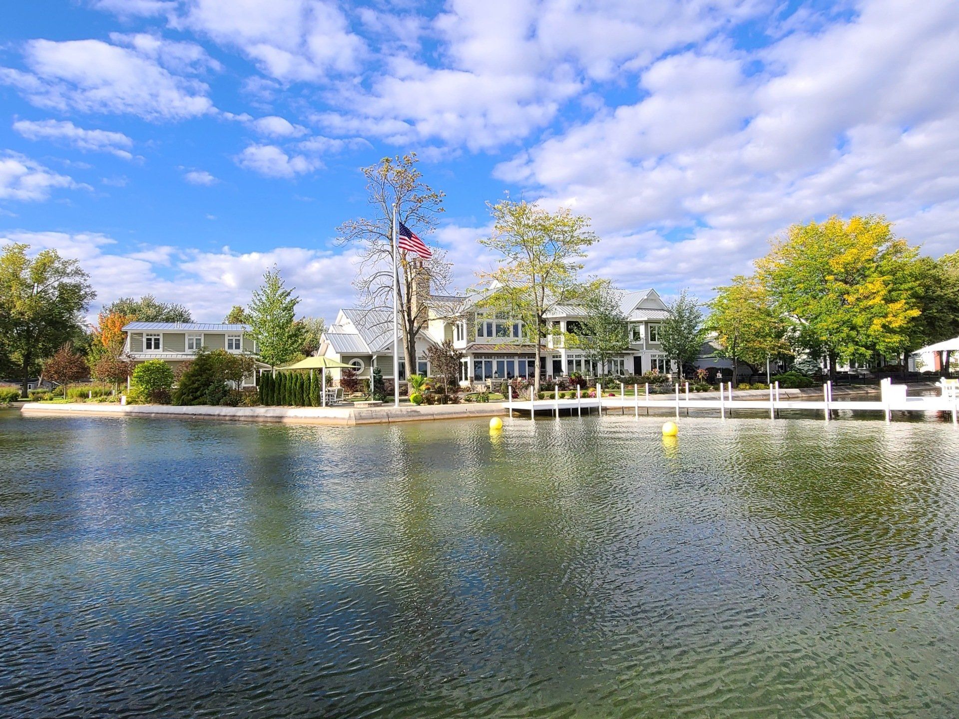 Lakeside house with a white facade and trees, reflected in calm water under a partly cloudy sky
