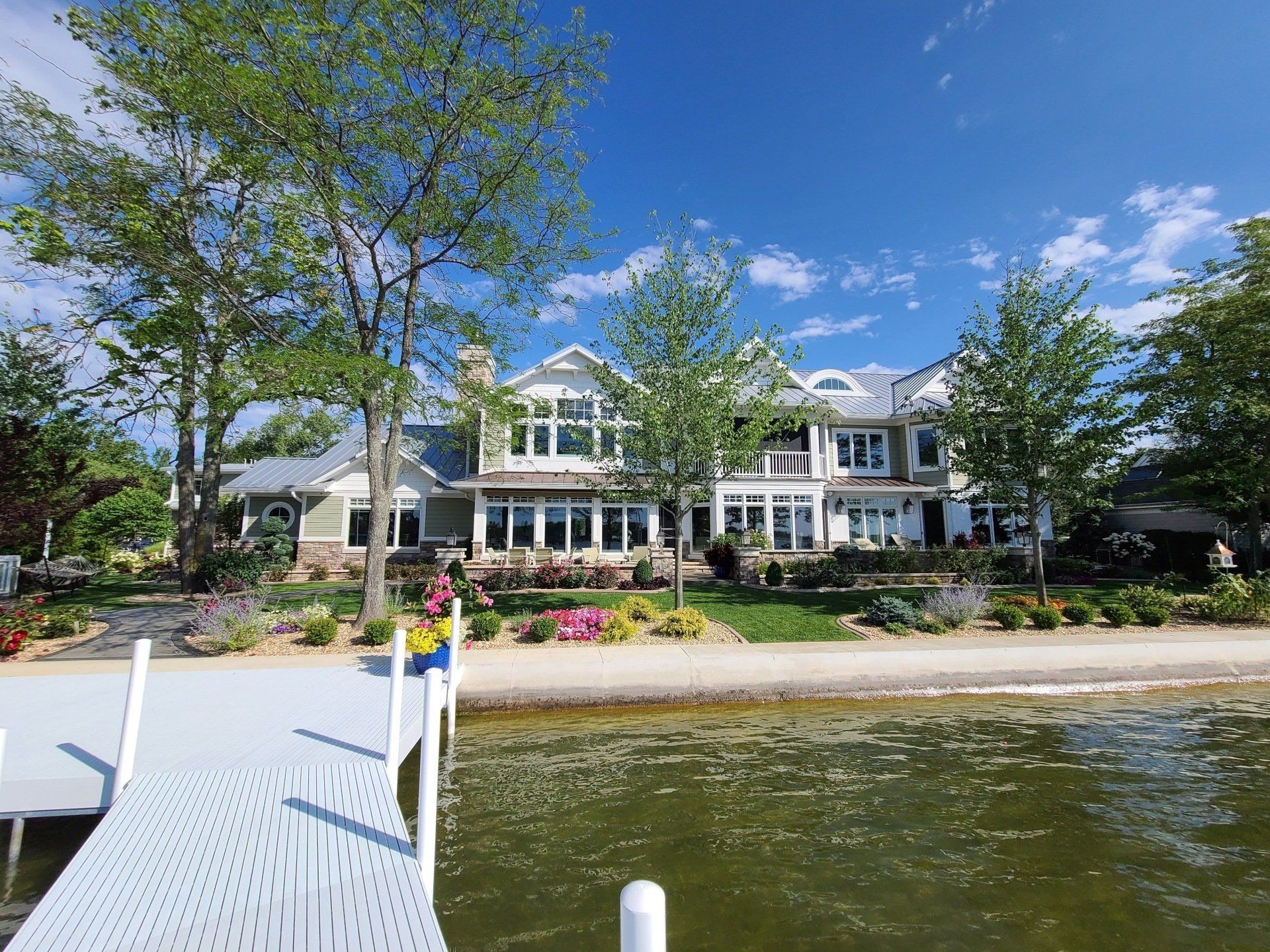 Lakeside mansion with dock in foreground, trees and flowers, under a bright blue sky.