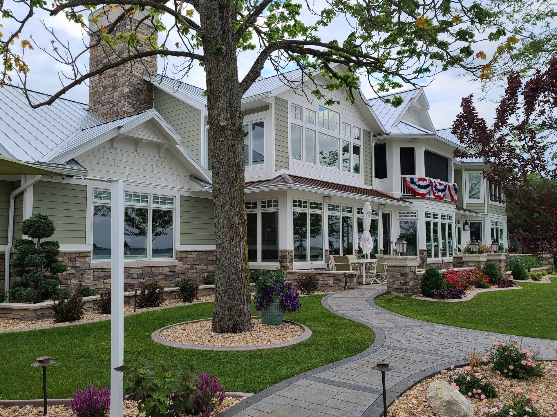 Large white Victorian-style house with wraparound porch, flower beds, and a curved stone walkway
