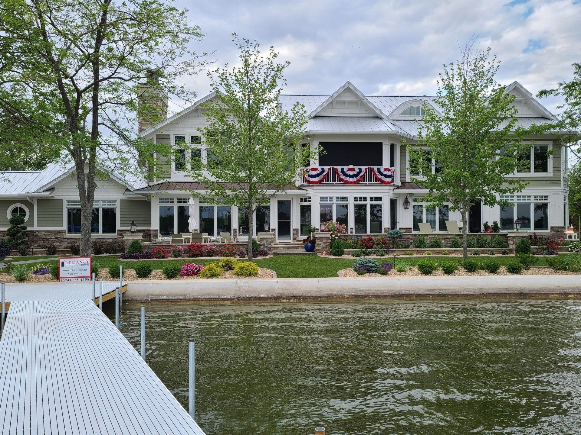 Large white lakeside house with patriotic bunting, trees, and a dock in the foreground