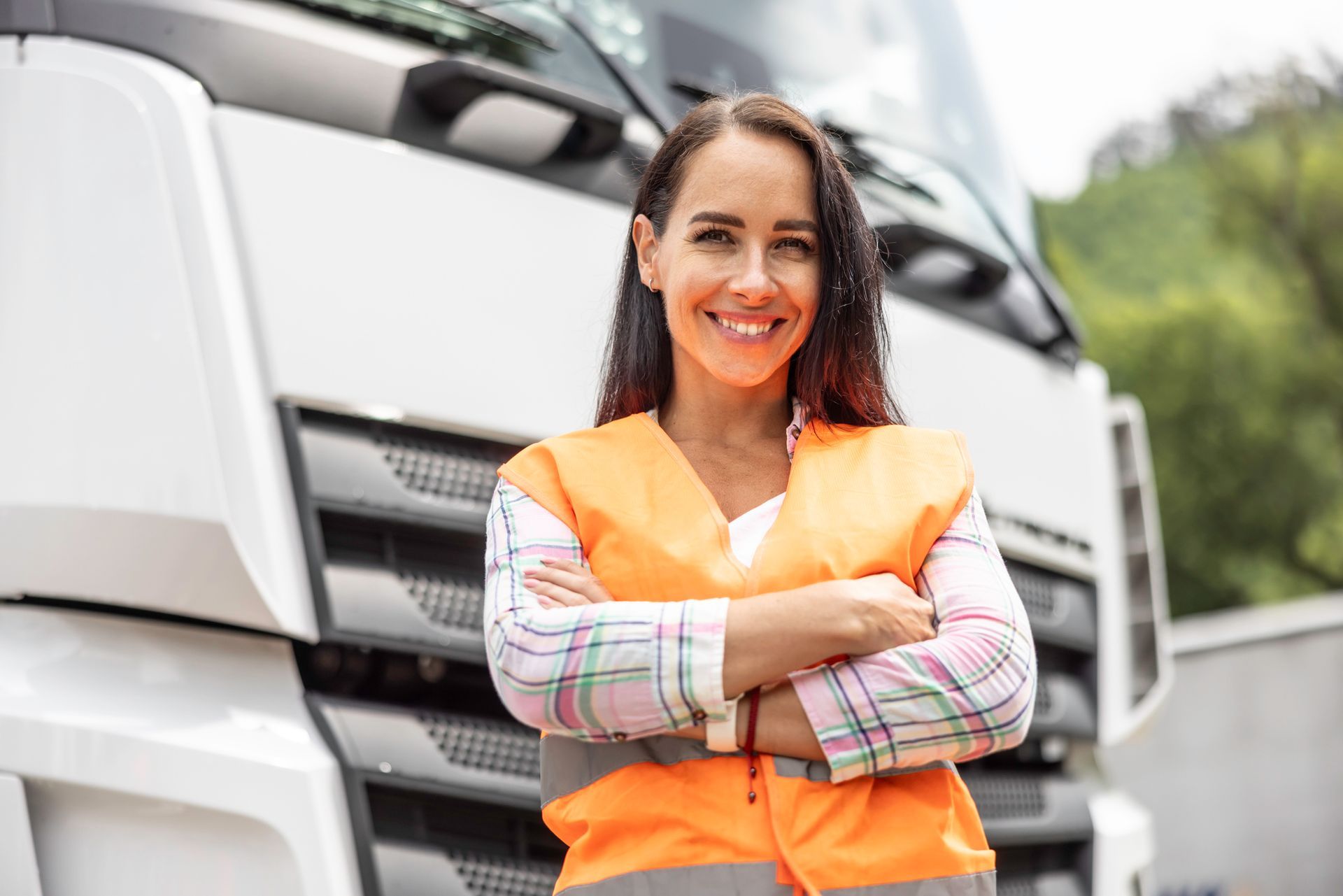 Smiling woman with arms crossed wears orange safety vest, standing in front of a semi truck Smiling woman with arms crossed wears orange safety vest, standing in front of a semi truck