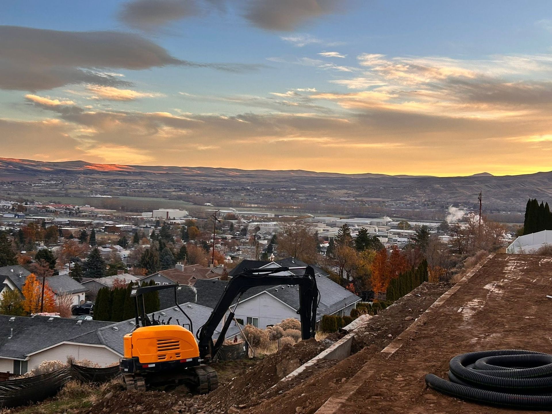 Excavator on a hillside overlooking a town at sunset with distant mountains and autumn trees.