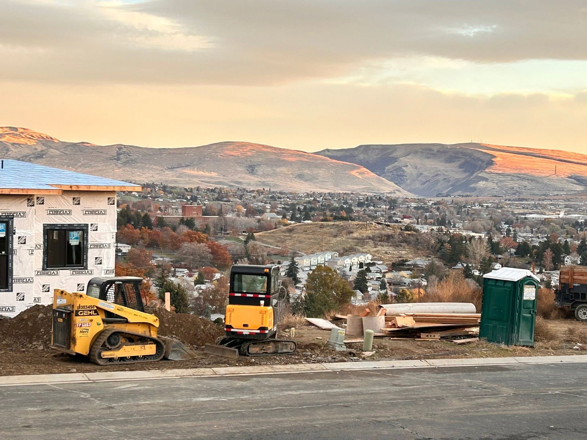Construction site with yellow loaders and containers overlooking a mountainous town at sunset