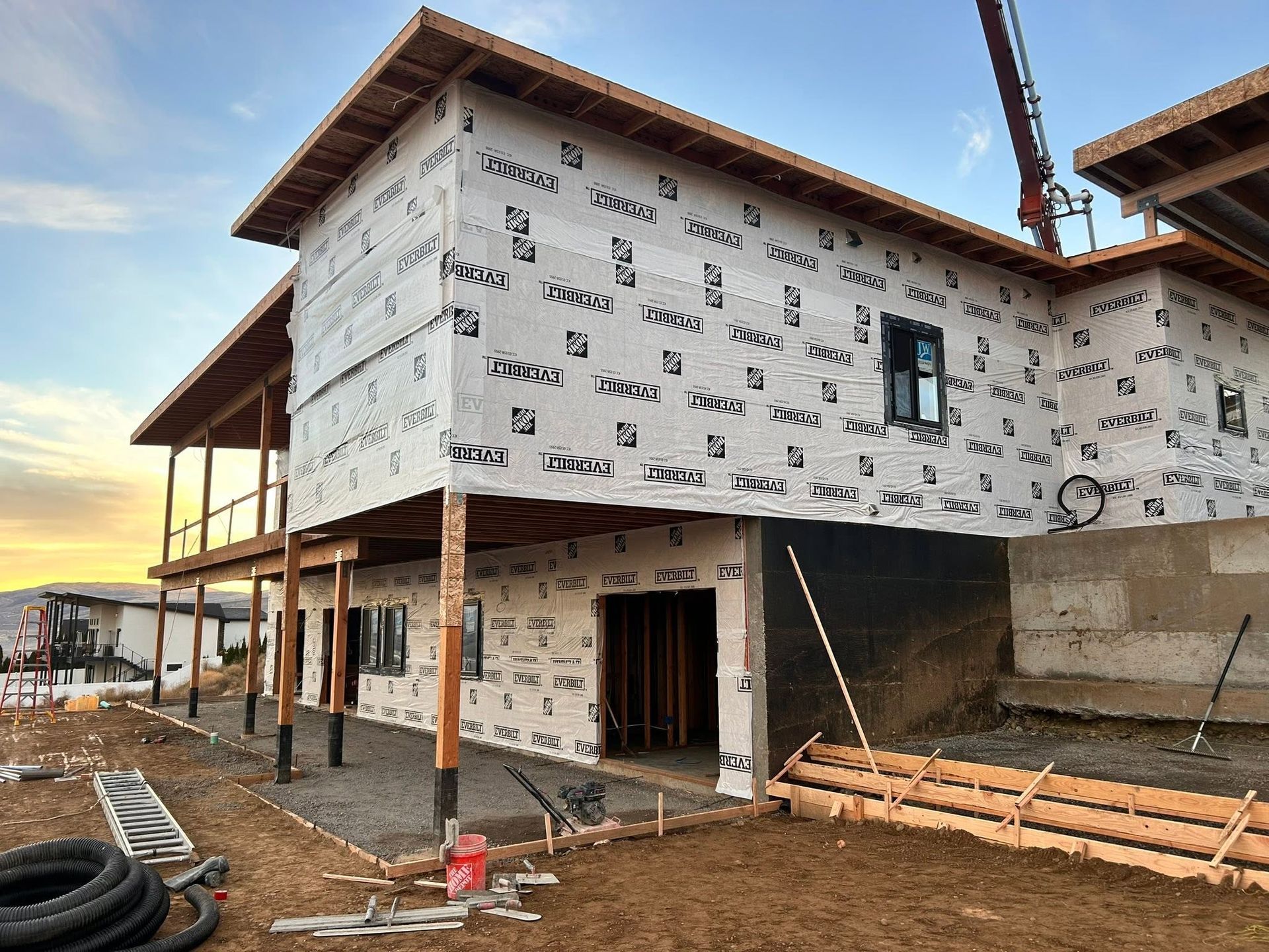 Partially built two-story house with white wrapped exterior, porch posts, and construction materials on muddy ground
