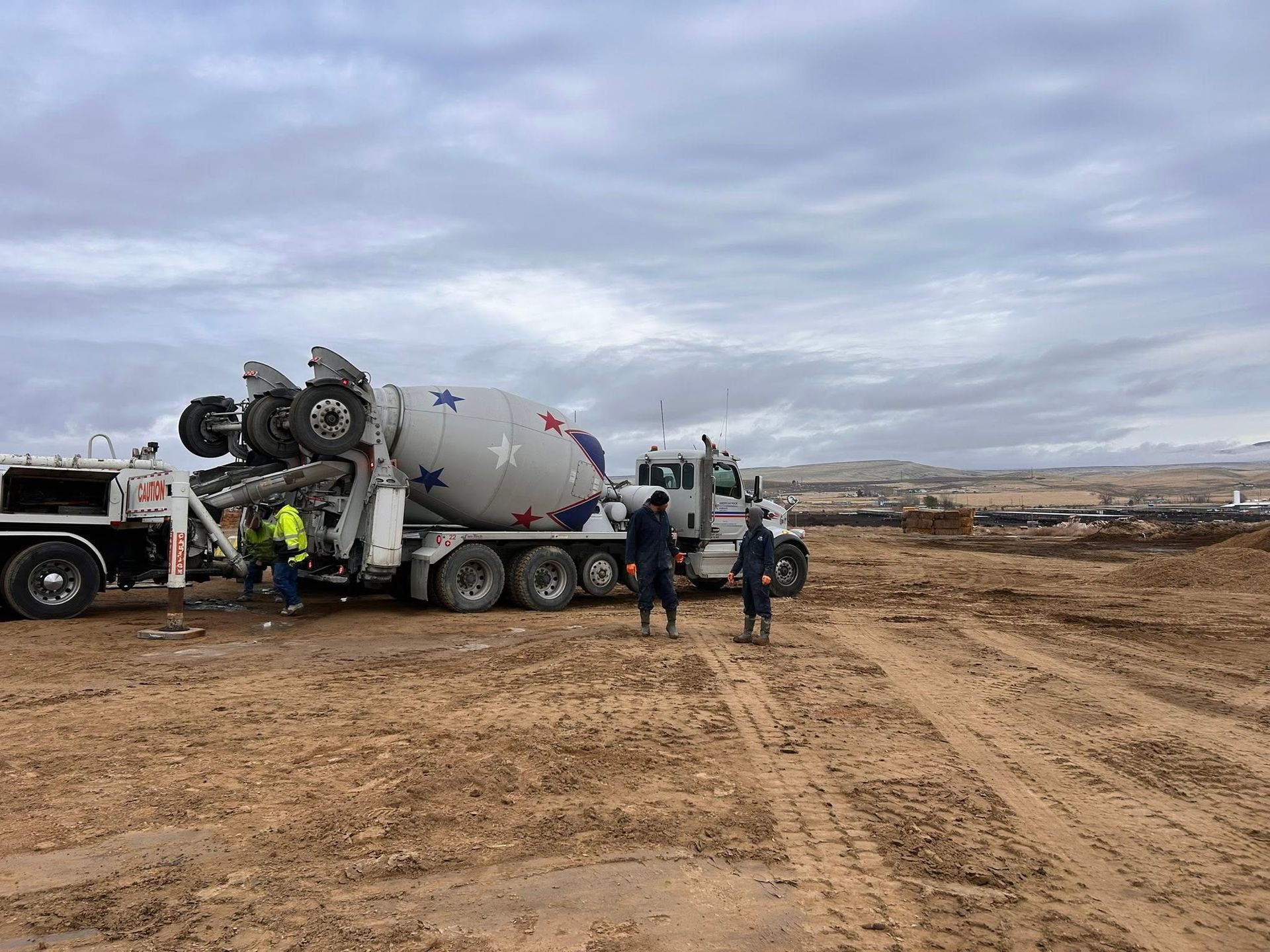 Concrete mixer truck and workers at a muddy construction site under a cloudy sky.