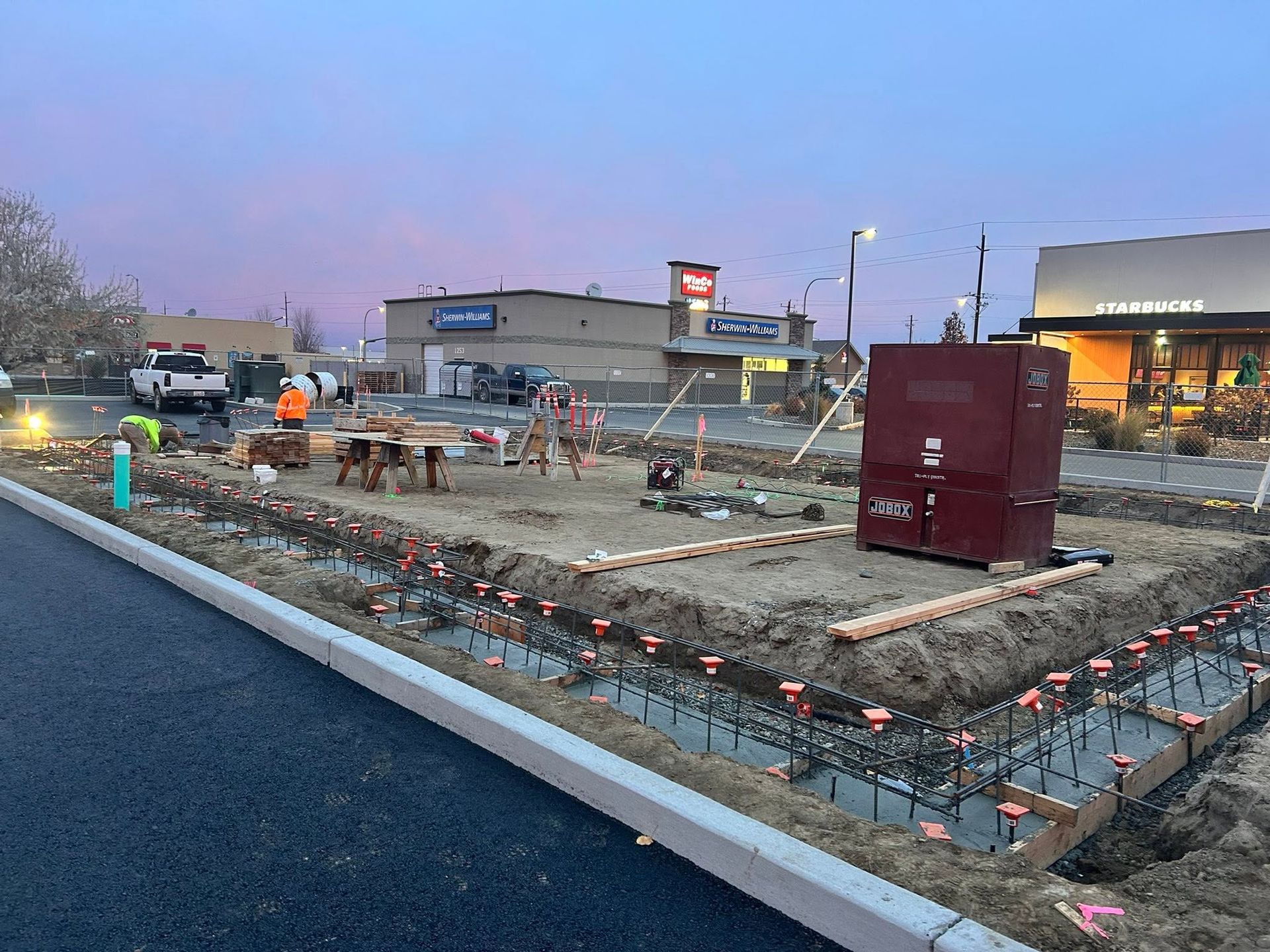 Construction site at dusk with concrete forms, rebar, orange lights, and a red dumpster beside a road
