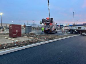 Construction truck with workers at dusk, pouring concrete along a paved road beside a curb and utility boxes
