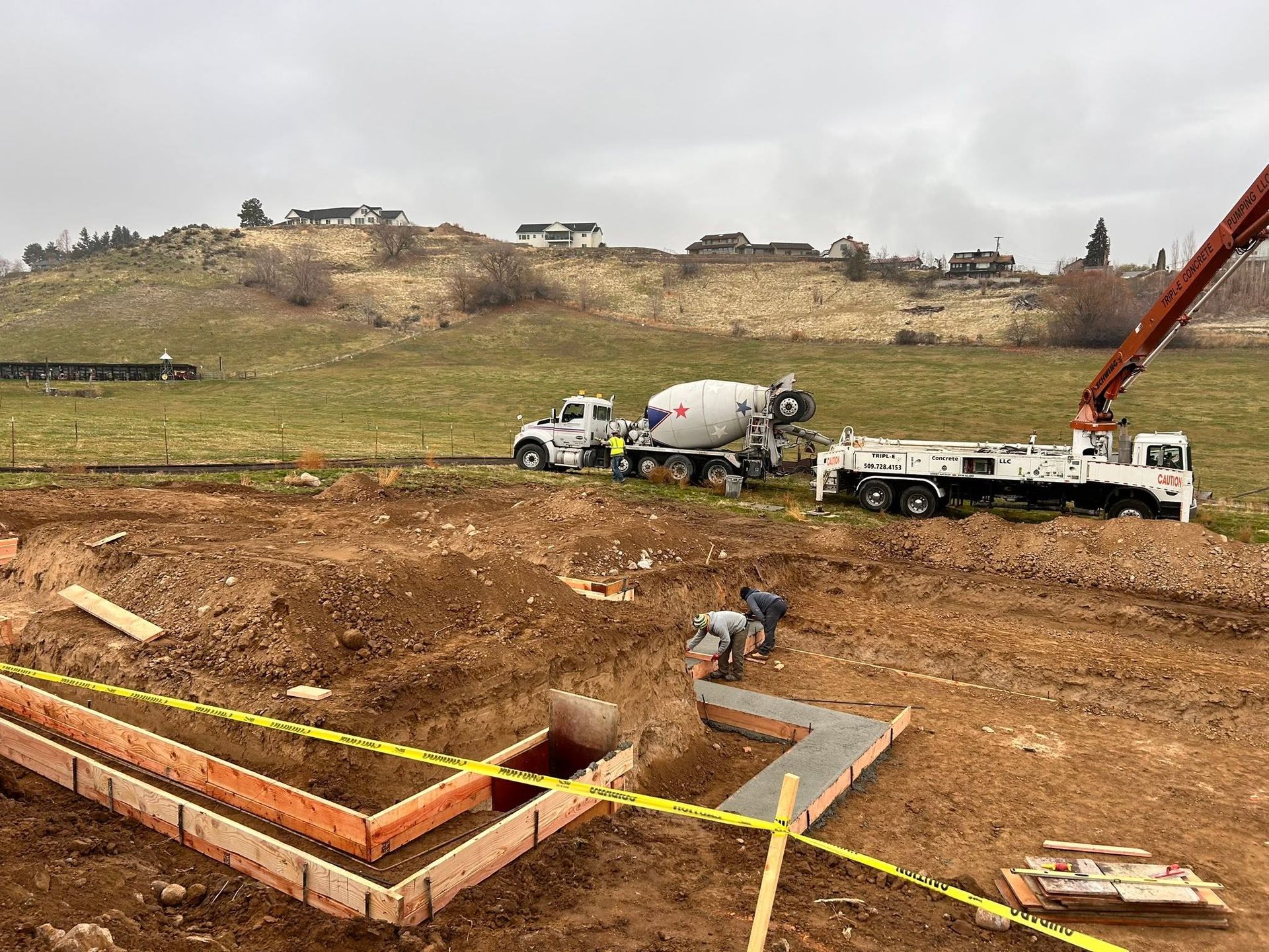 Construction site with concrete foundation, workers, cement truck, and excavator on a dirt hillside