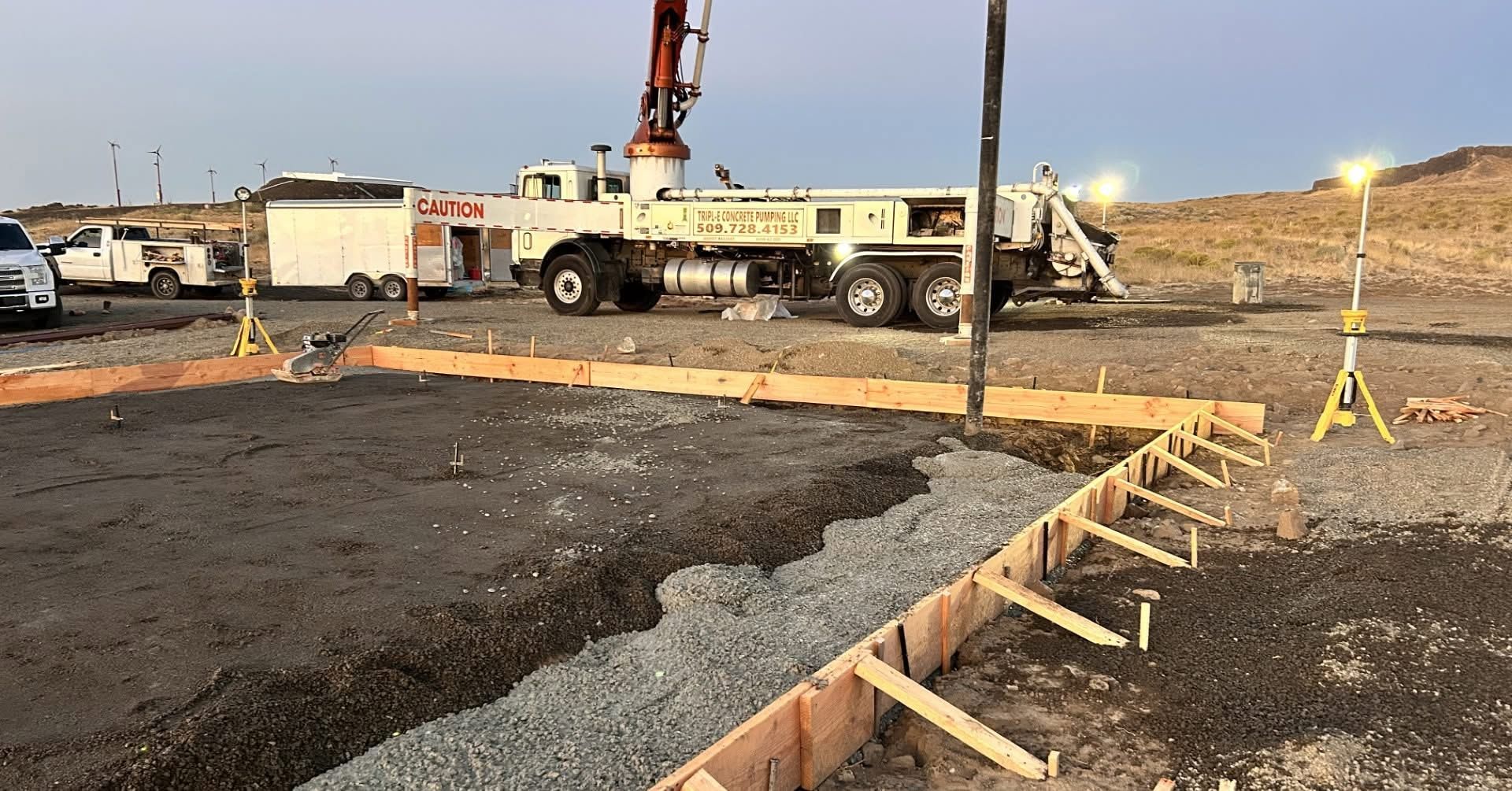 Construction site with trucks, crane, and workers paving a dark asphalt area alongside a dirt road.