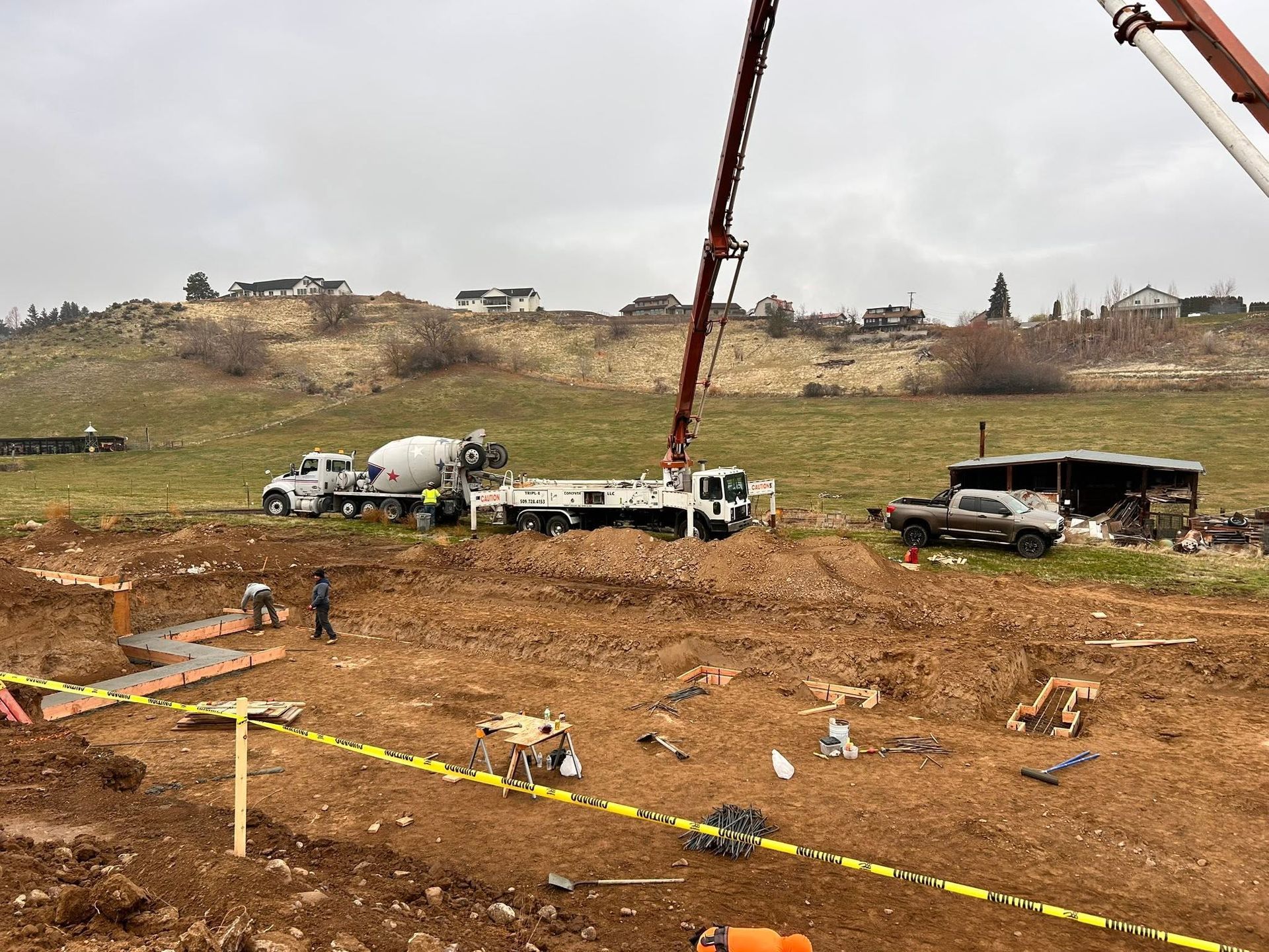 Construction site with cement truck and crane pouring concrete into an excavated lot, with yellow caution tape.