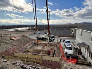 Construction site with concrete foundation forms, crane, trucks, and a partially built gray building under a cloudy sky
