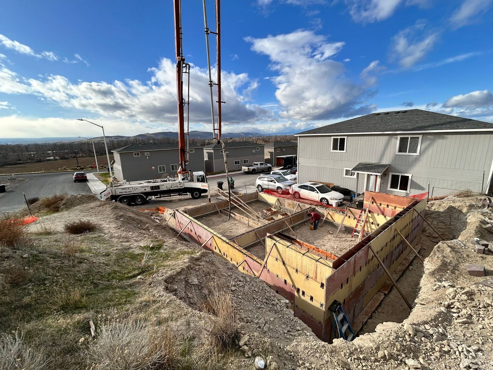 Construction site with a foundation pit, red crane, and a gray house under a blue sky