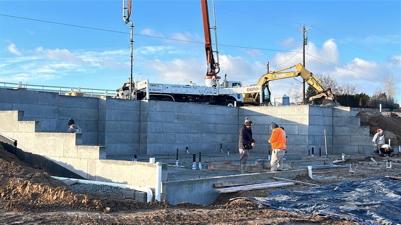 Concrete retaining wall under construction with workers, excavator, and crane on a snowy site