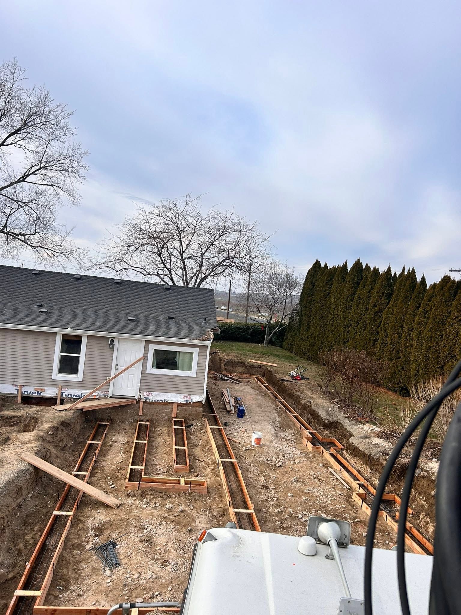 Backyard under construction beside a gray house, with wooden framing, gravel, and tall hedges.