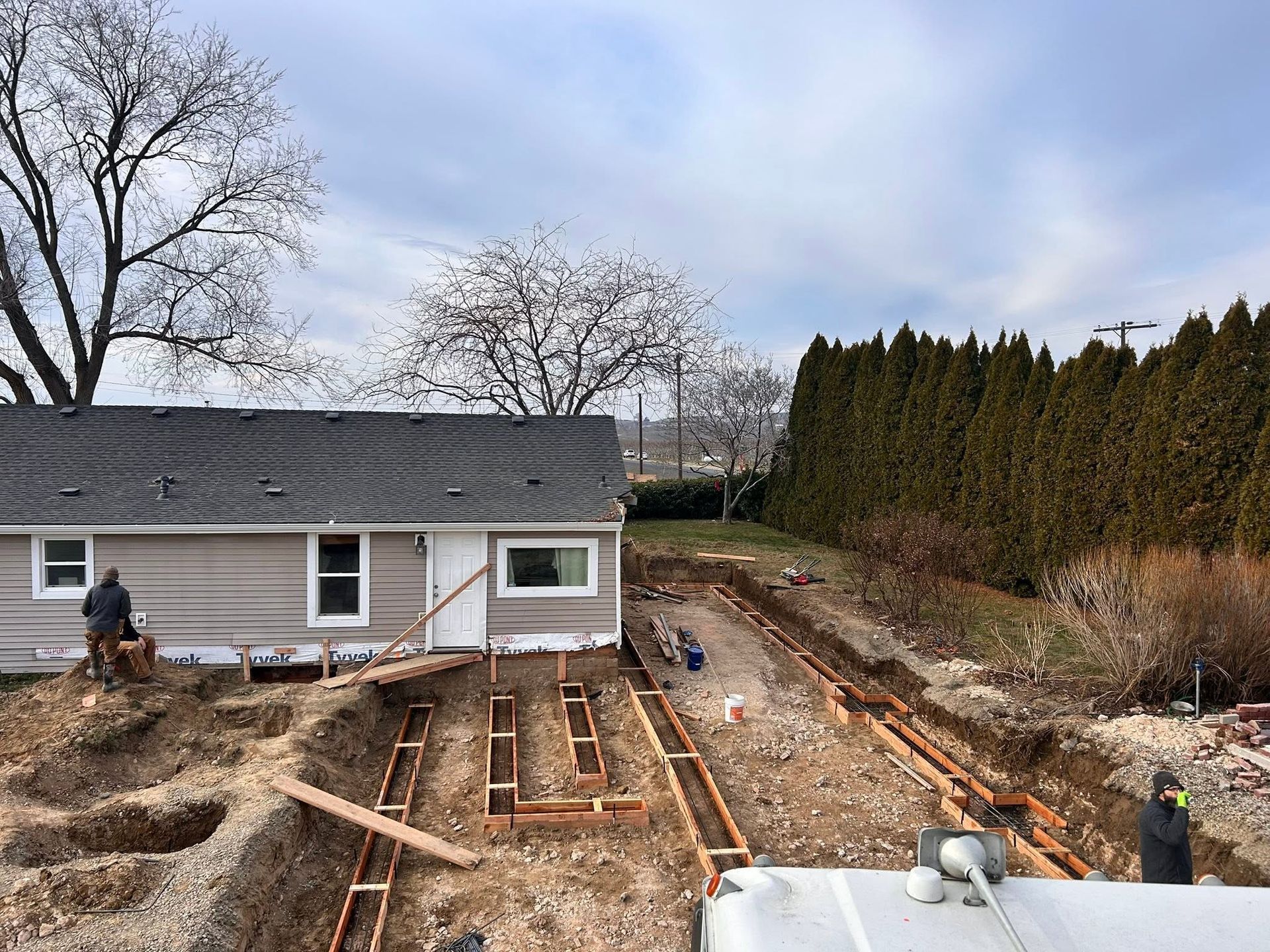 Backyard construction trench beside a house, with workers and wood framing under cloudy sky
