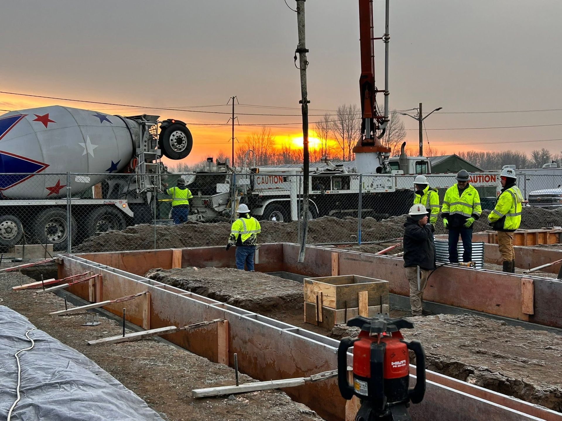 Construction crew working on a concrete foundation at sunrise beside a cement truck and excavator.