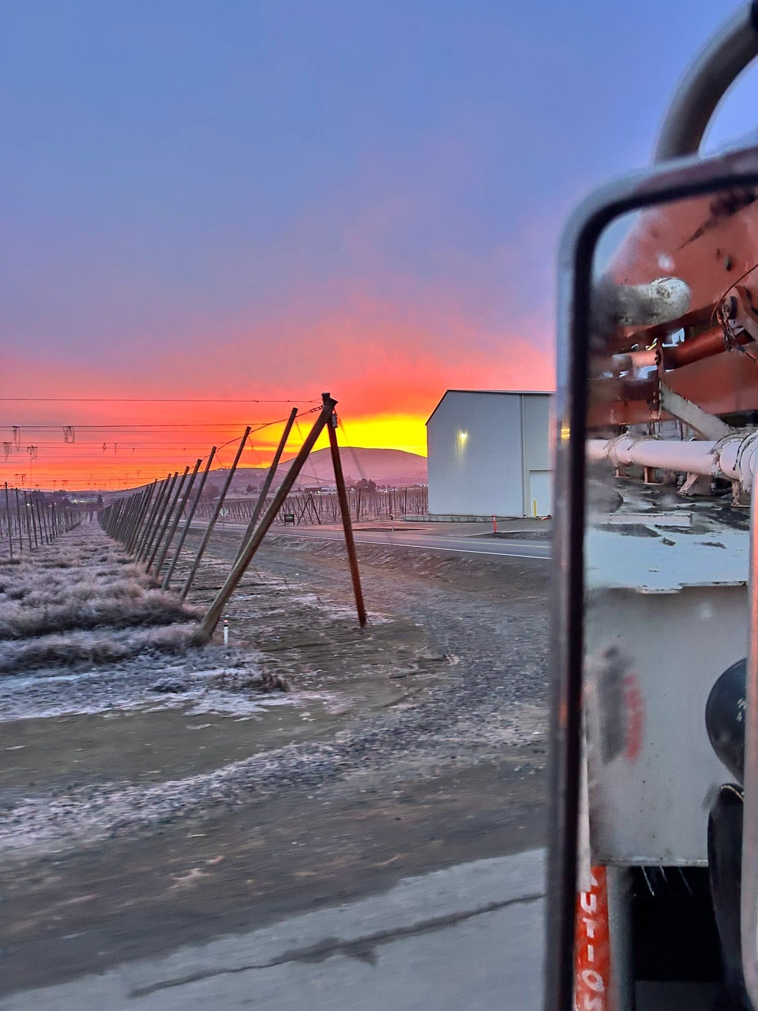 Snowy roadside at sunrise with red-orange sky, fence, and a vehicle side mirror reflecting machinery.