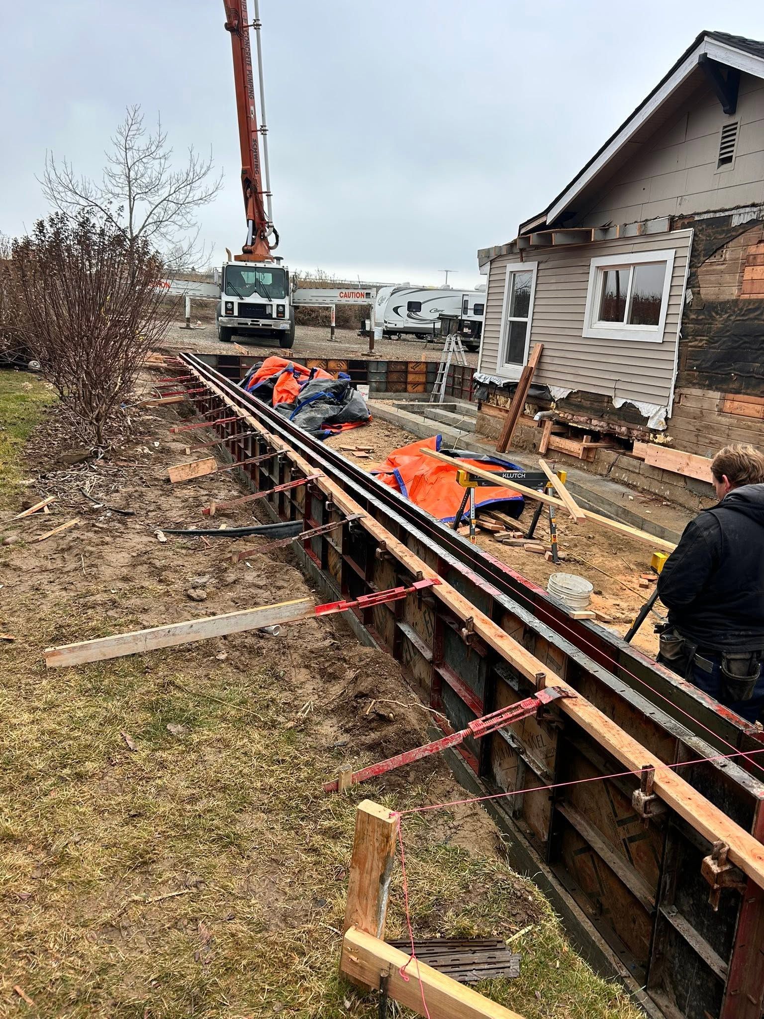 Trench excavation beside a house, with workers, pipes, and an excavator on an overcast day