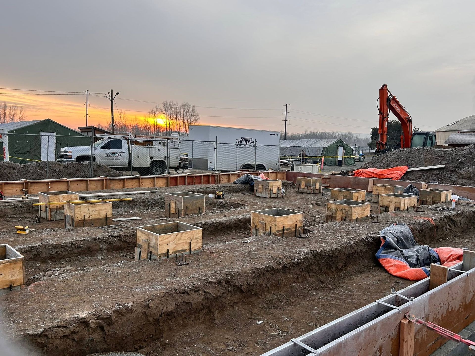 Construction site with trenches, wooden forms, excavator, and vehicles at sunset