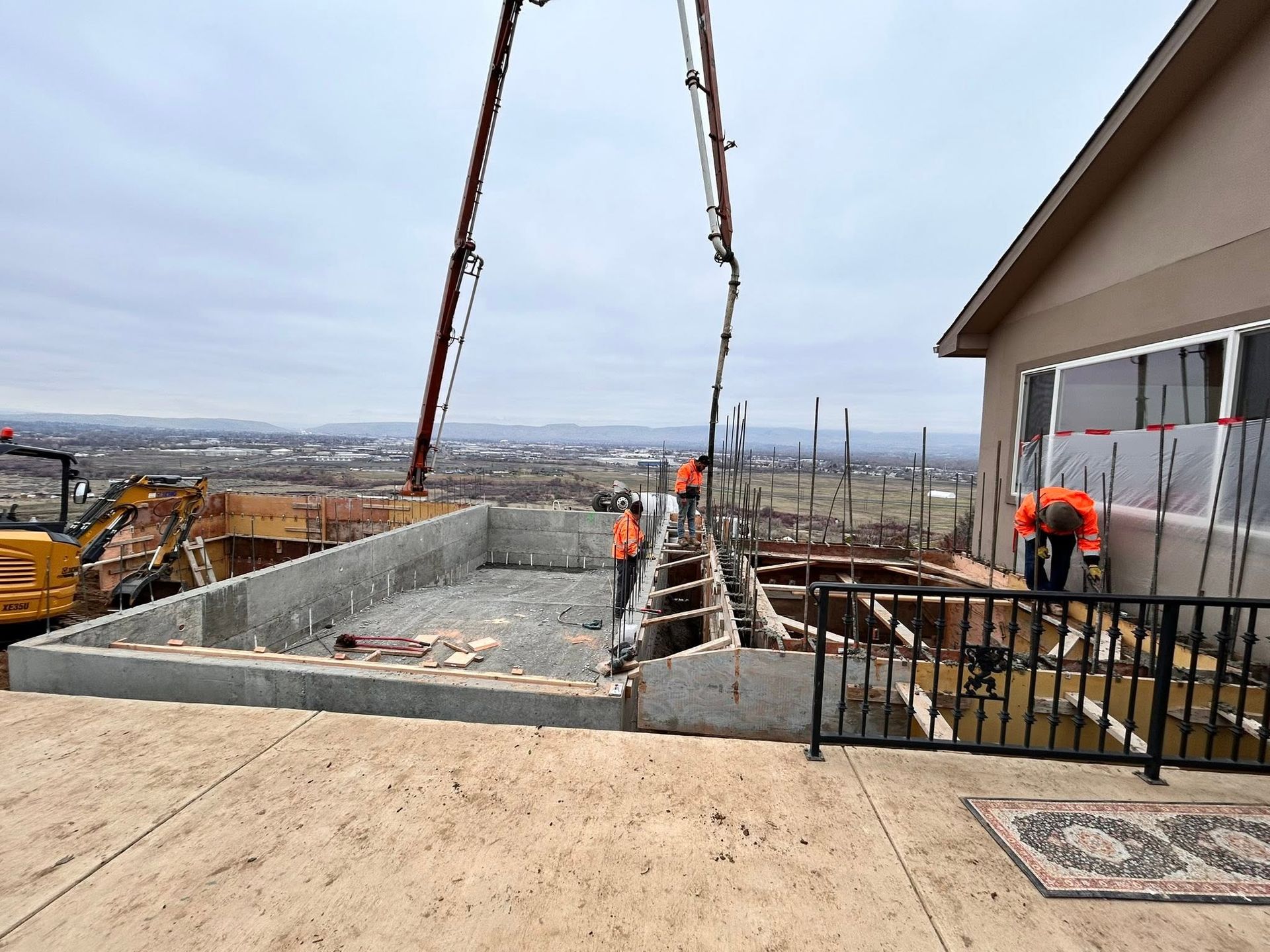 Construction site with workers, crane cables, and concrete foundation beside a building.