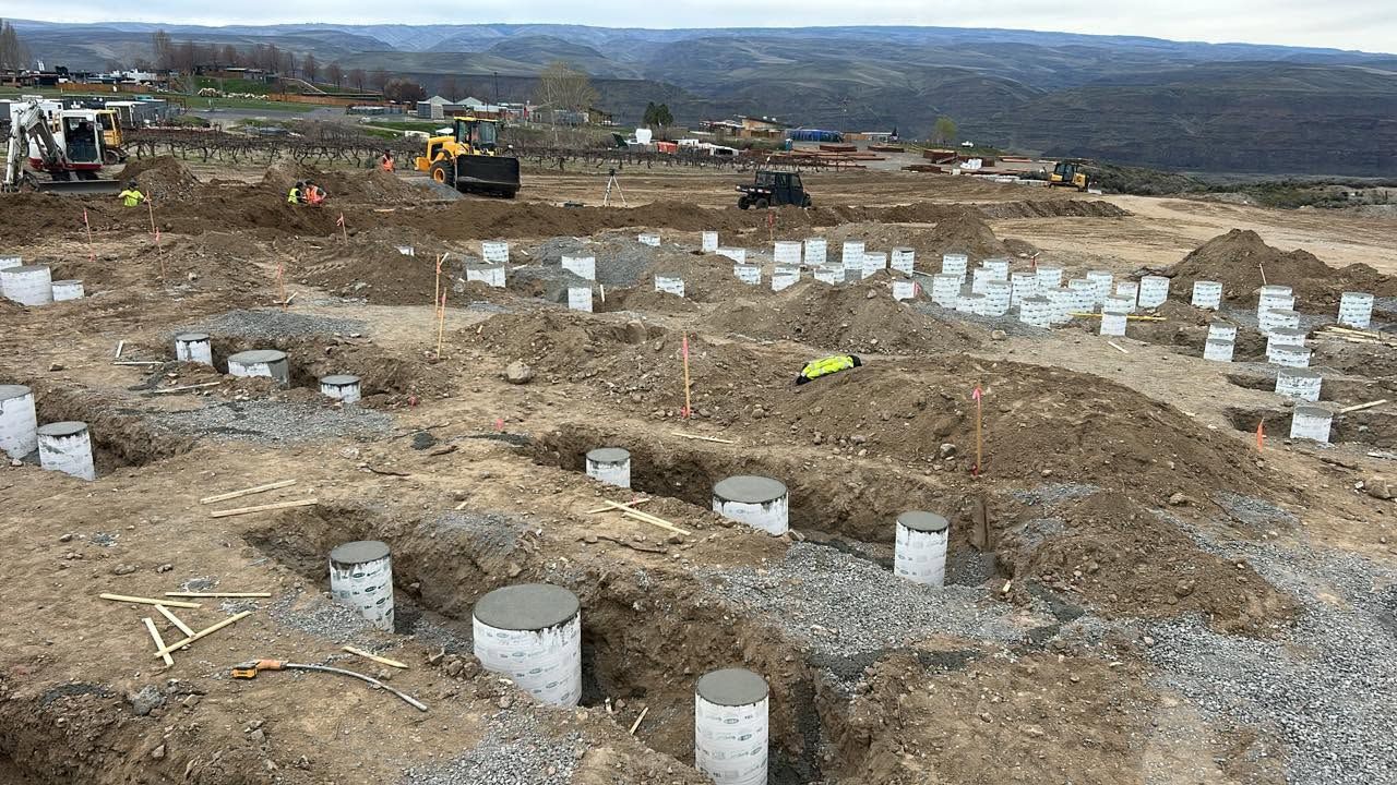 Construction site with concrete foundation footings and excavators on a dirt hillside under cloudy skies