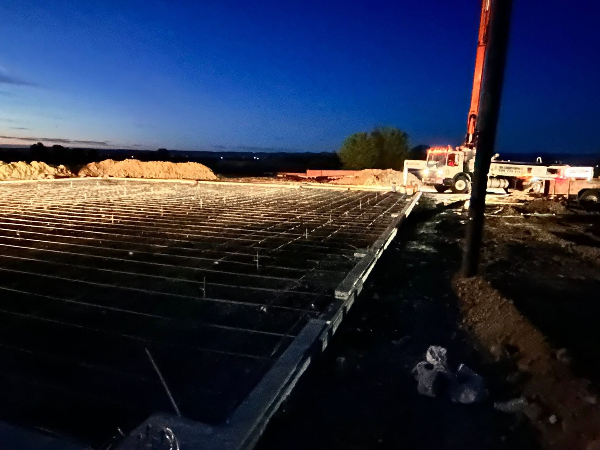 Nighttime construction site with rebar grid, dirt piles, and a lit building under a dark blue sky