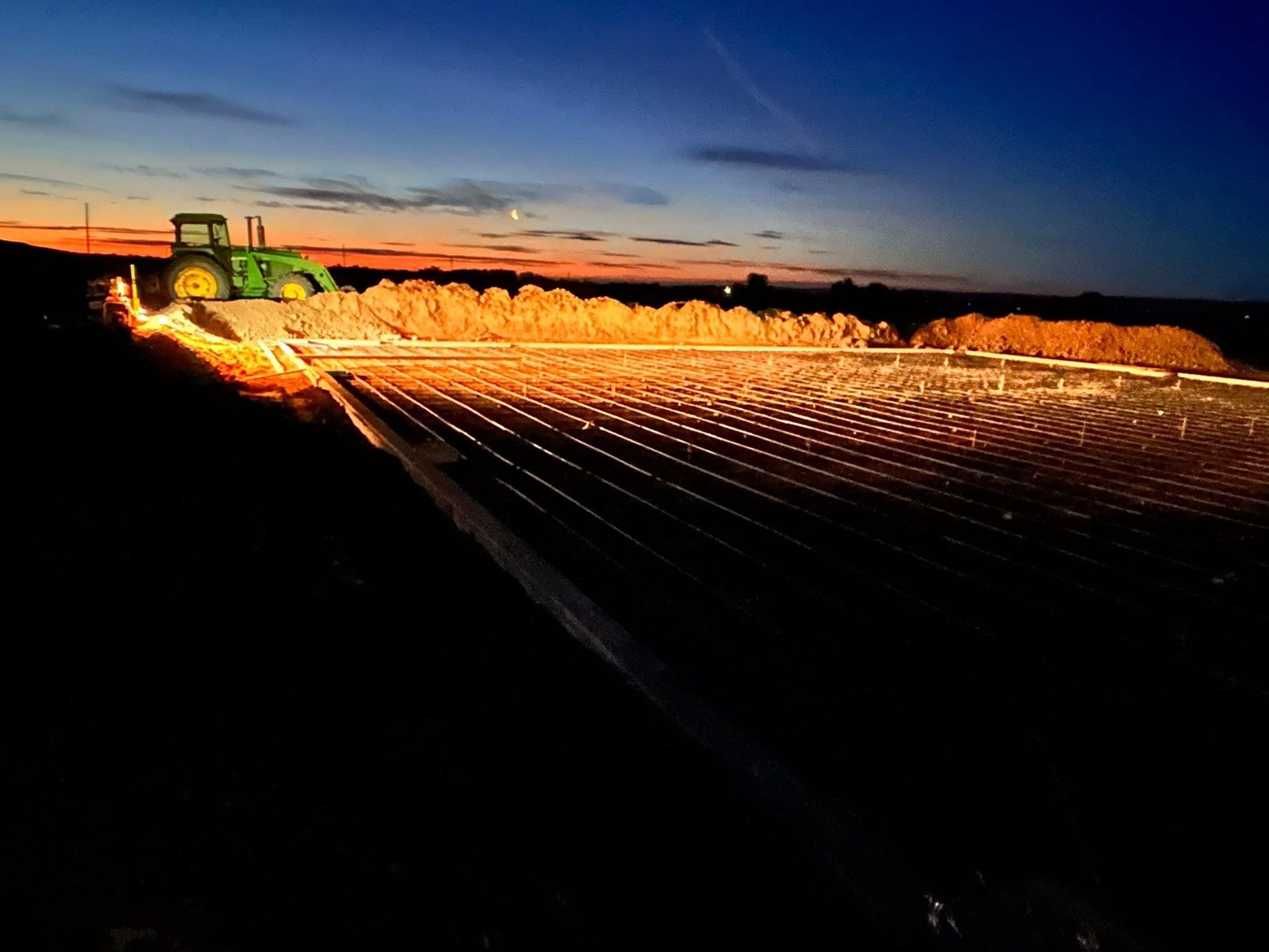 Tractor harvesting a field at sunset, with bright lights illuminating rows of crops.