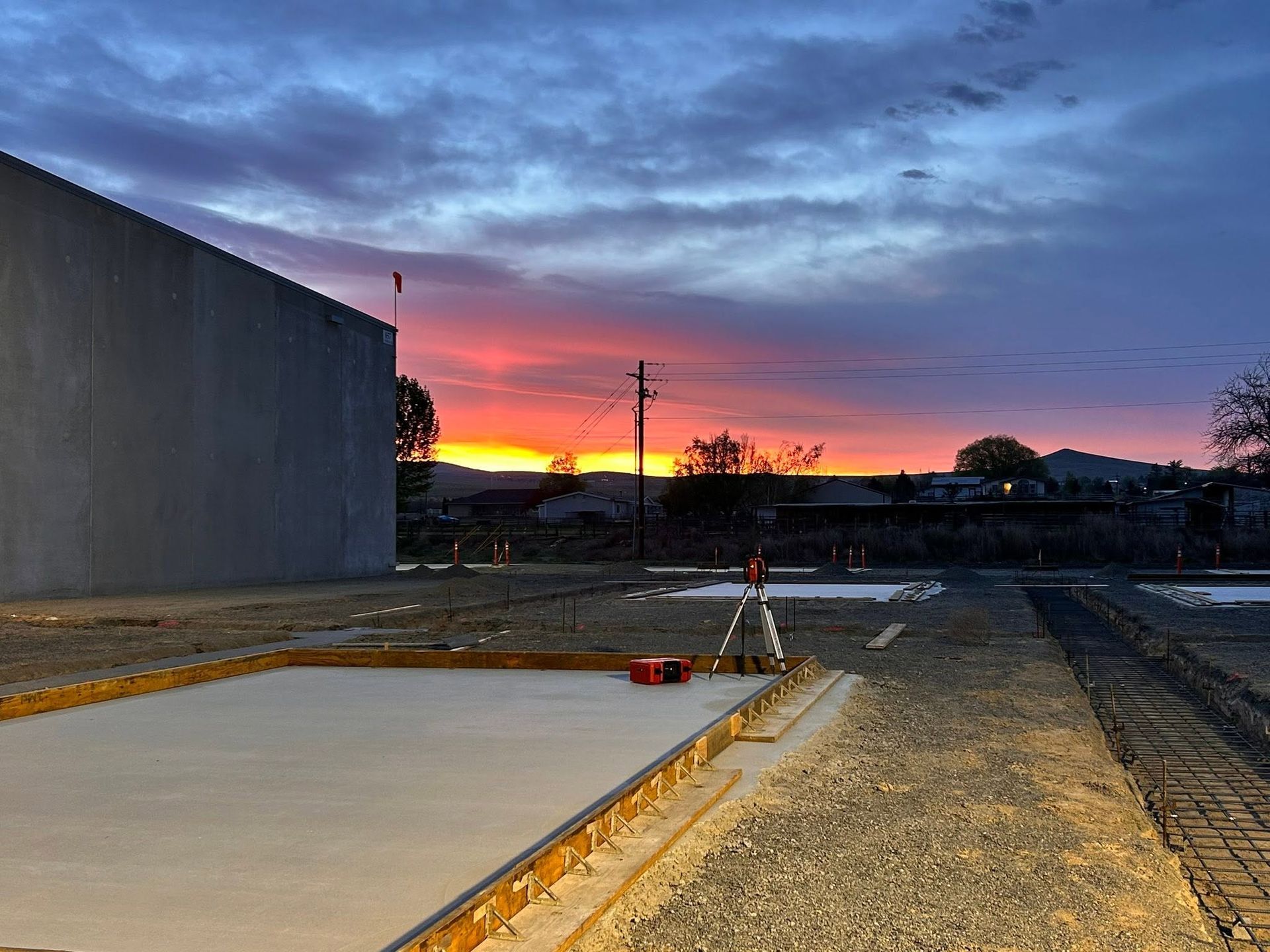 Empty construction lot at dusk with a sunset sky, concrete foundation, and gravel road beside a large building.