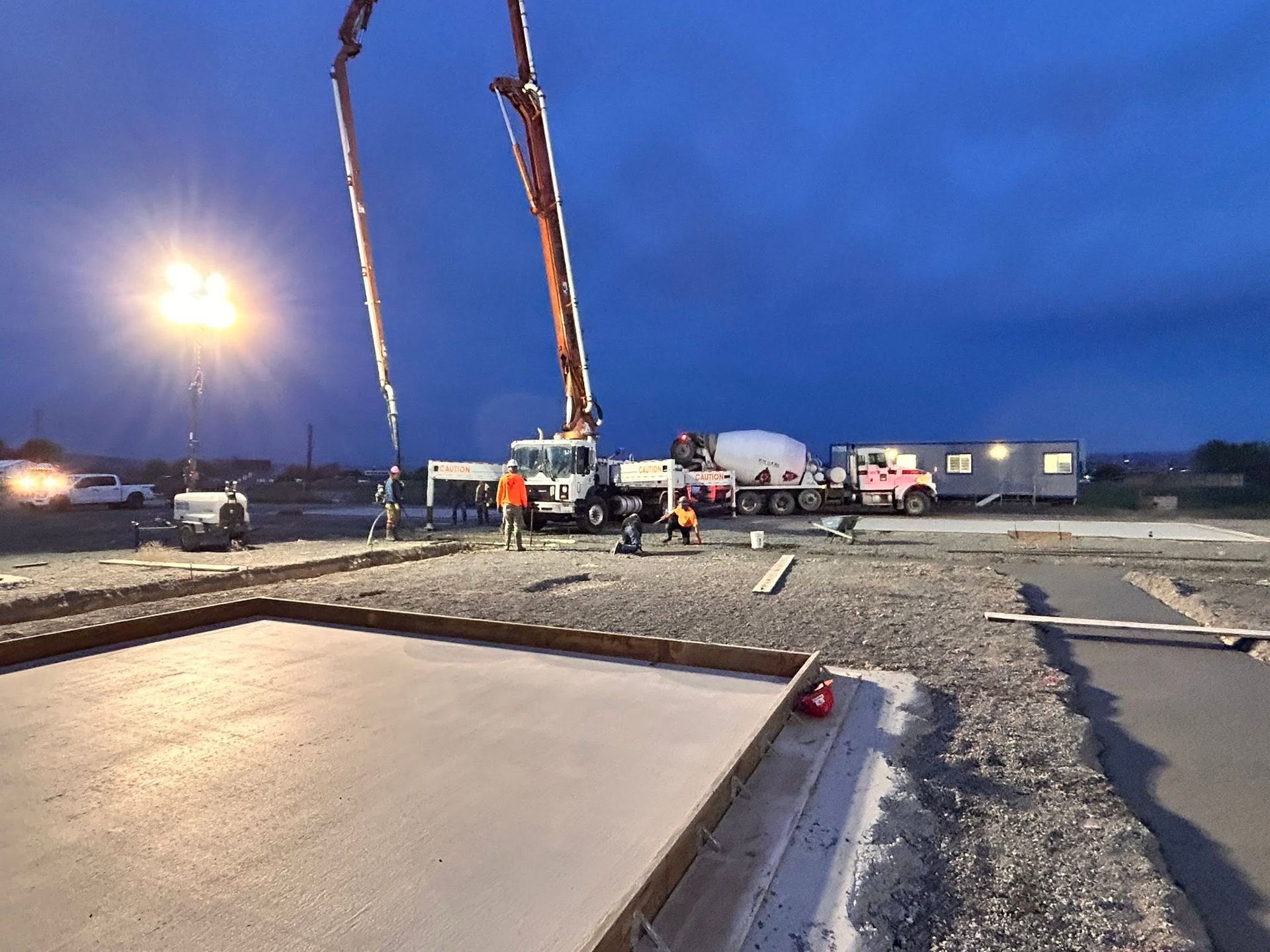 Concrete pump truck pouring at a construction site at dusk, with workers and equipment under bright lights