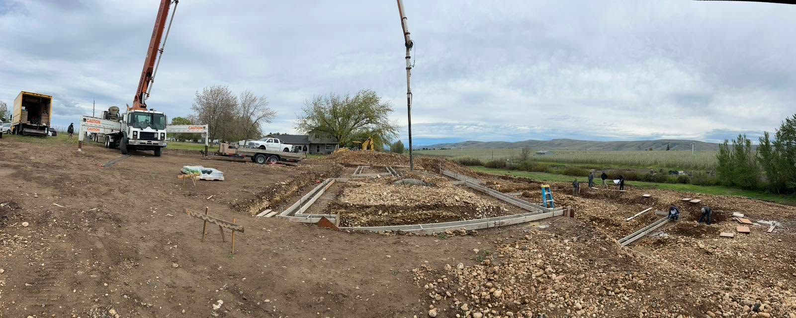 Construction site with crane, trucks, and rebar foundation on a dirt hillside under cloudy skies