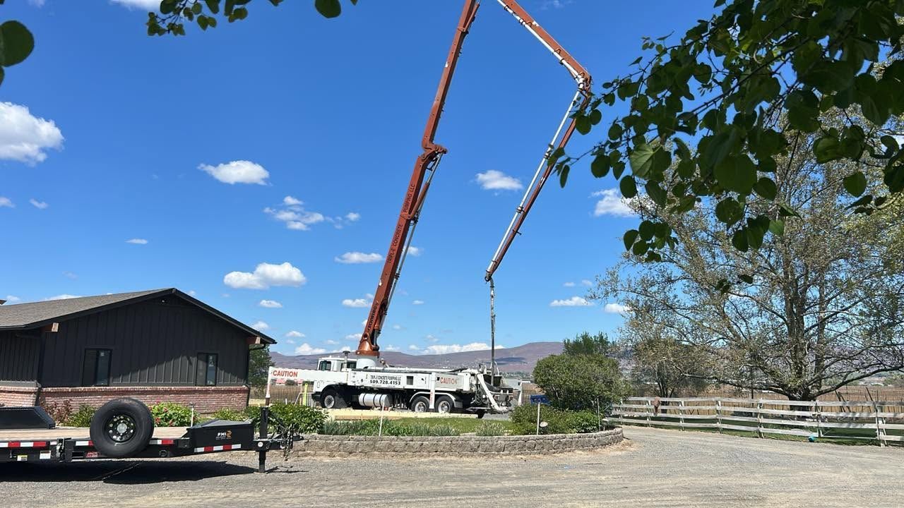 Two orange concrete pump booms stretch over a construction site beside a dark barn under a blue sky.
