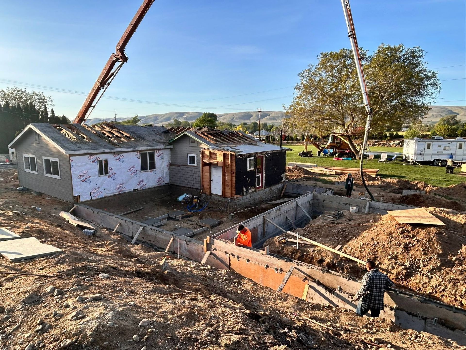 House under construction with cranes lifting materials over a dirt foundation on a rural property.