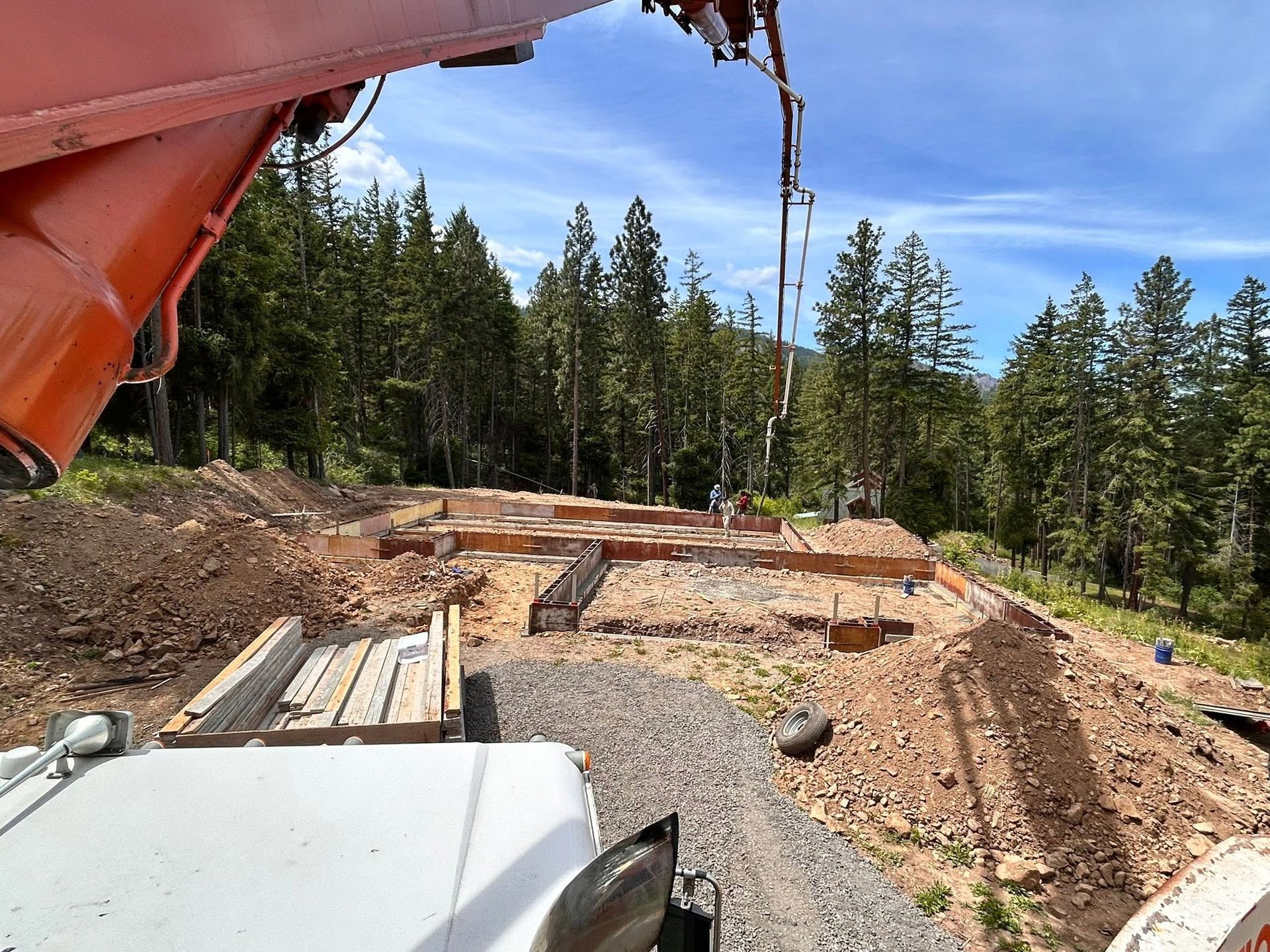 Rural construction site with gravel pads, lumber, and a muddy excavator bucket against a forest backdrop