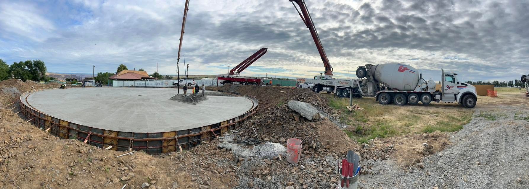 Construction site with a large concrete foundation slab, crane, cement mixer truck, and gravel under a cloudy sky