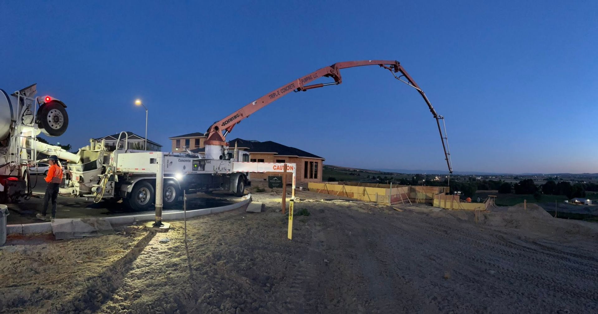Concrete pump truck pouring at a construction site at dusk, with workers and houses in the background