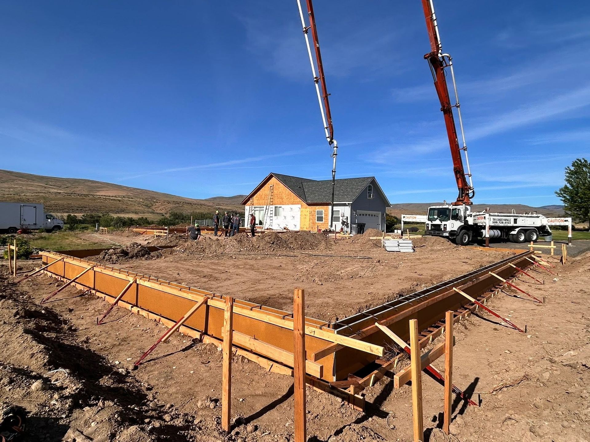 Concrete foundation formwork at a rural house site with two crane booms overhead.