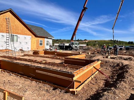 Construction site with concrete foundation forms, crane, and workers beside a brick building under a blue sky