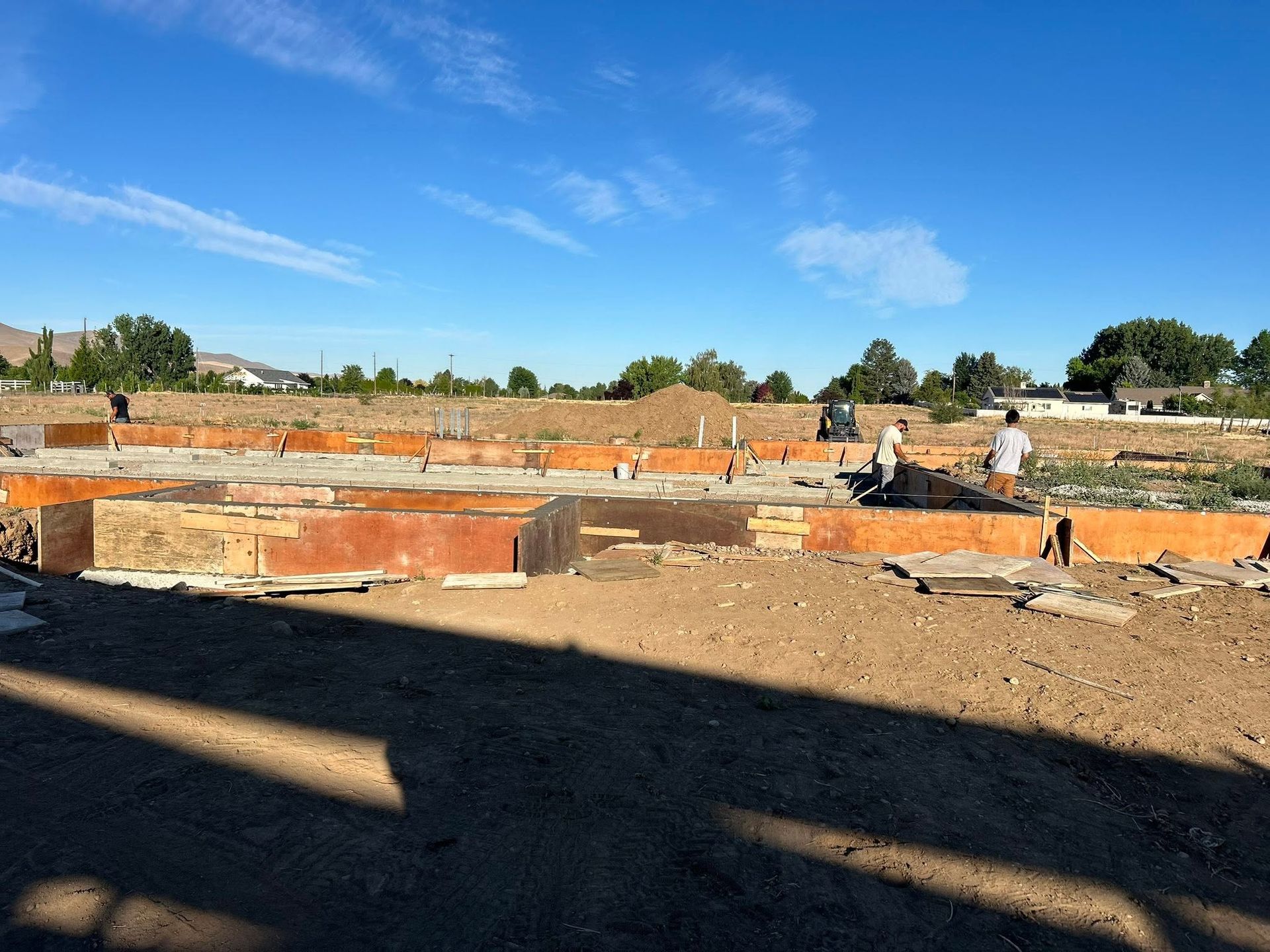 Construction site with orange foundation walls, dirt foreground, and workers under a blue sky