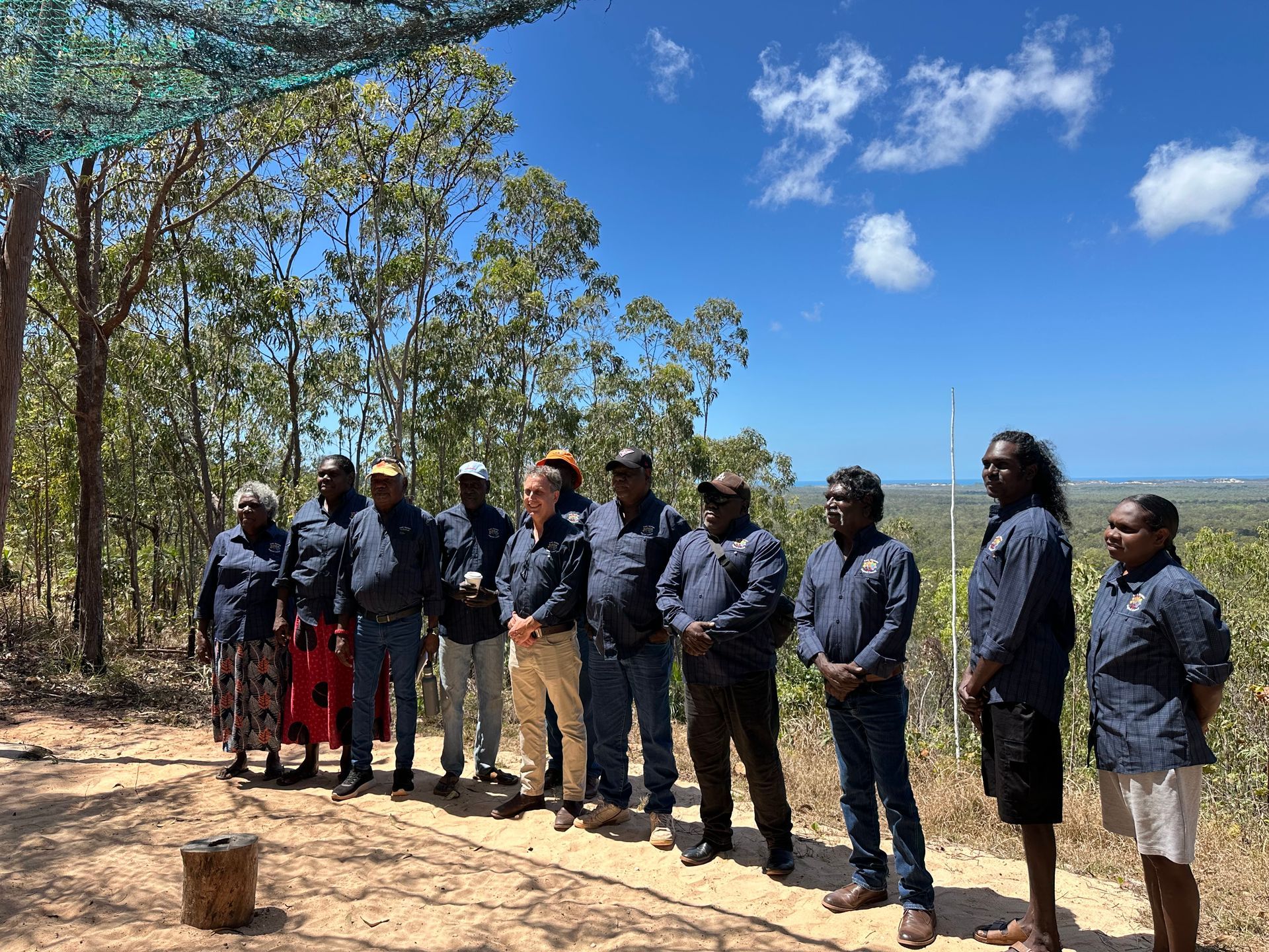 A group of people standing together. In the background are trees and blue sky. 