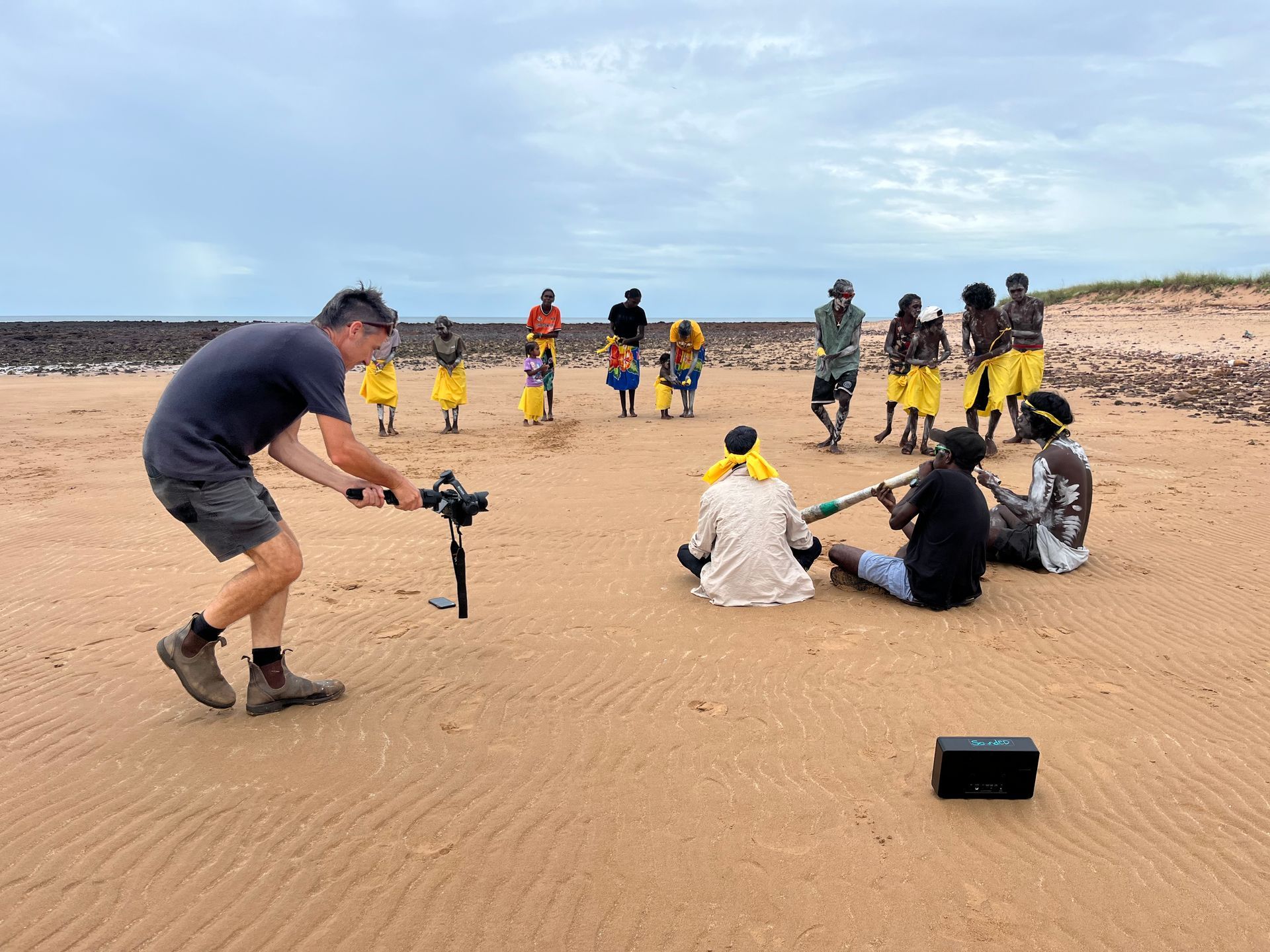 A man films a group of people singing and dancing on the beach.