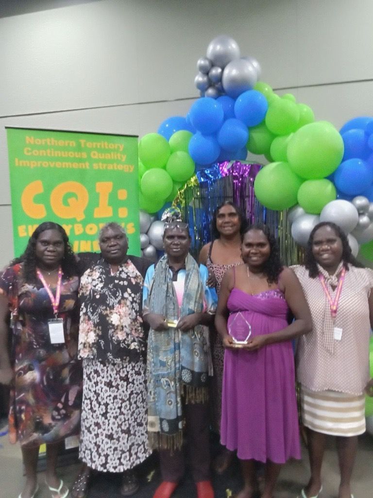Six women standing together looking happy. Two are holding an award.