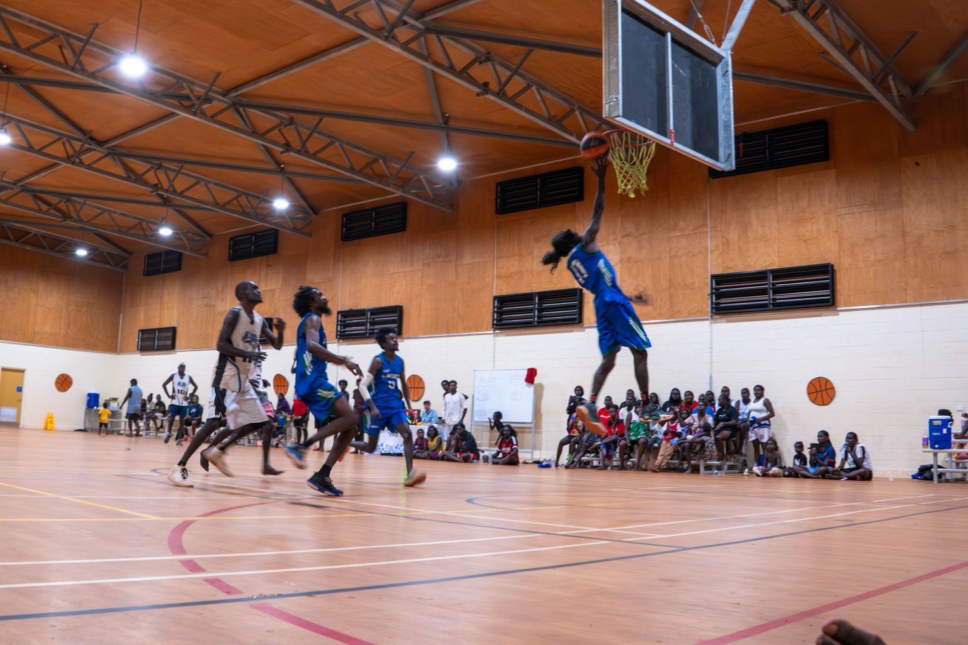 A team of men playing basketball. One player jumps in the air near the hoop to score. Spectators are sitting and standing on the sideline.