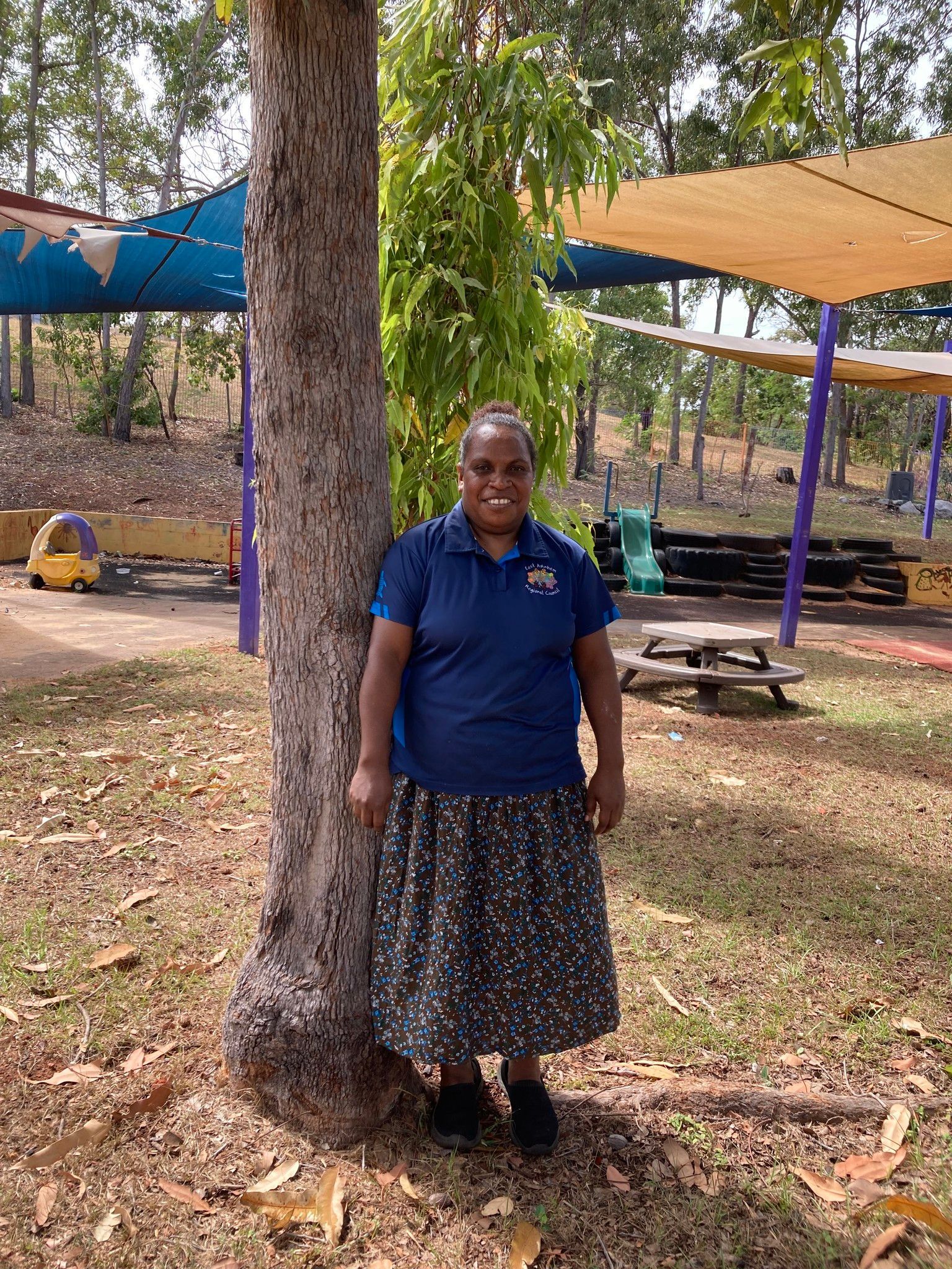 A woman standing and smiling. An outdoor playgroup is in the background. 