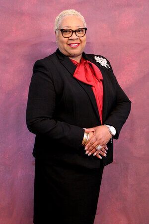 Portrait of a smiling person in a black suit with a red blouse, standing against a pink backdrop.