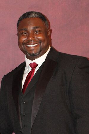 Smiling man in a black suit, white shirt, and red tie against a pink backdrop