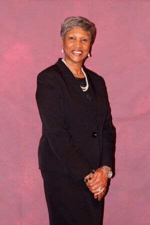 Person in a black suit smiling against a pink studio backdrop, hands clasped.