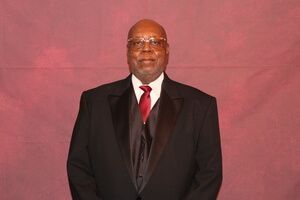 Man in a black suit, white shirt, and red tie standing against a plain pink backdrop.